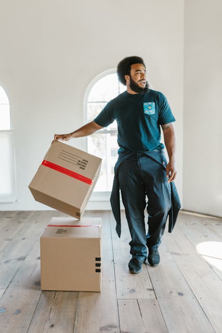 A male mover with dark skin and curly hair, dressed in a navy blue t-shirt with a logo on the chest, is inside a bright, empty room with white walls and wooden flooring, engaged in a home relocation process. He is holding a medium-sized cardboard box wrapped with red tape in his right hand, preparing to load it onto a larger box placed on the floor. The room features a large arched window allowing natural light to fill the space, indicating daytime. In the background, there are no additional furniture or objects visible, emphasizing the focus on packing and moving activities. The scene captures the logistics and muscle involved in furniture transport and packing during a house removal, consistent with professional moving services provided by Self Storage Pimlico.