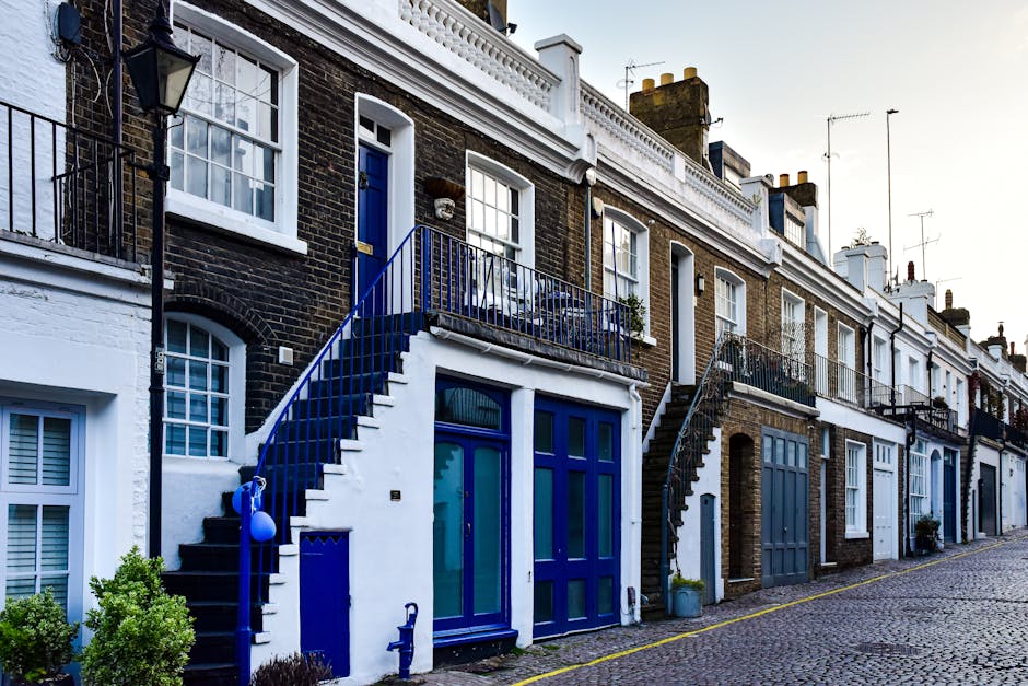 A row of Victorian-style terraced houses with ornate white facades and large bay windows facing a narrow street in Pimlico, London. Several parked cars, including a silver hatchback in the foreground and various other vehicles in different colors, are lined along the curb. The street is lined with mature trees providing partial shade, and a hotel sign is visible on one building. The pavement is clean, and the scene is captured during daylight with overcast skies, indicating typical urban residential surroundings suitable for house removals and furniture transport. The image reflects a typical residential area where Self Storage Pimlico conducts home relocation and packing and moving services, with the street activity and environment supporting efficient loading and unloading processes for small flat moves in the Pimlico area.