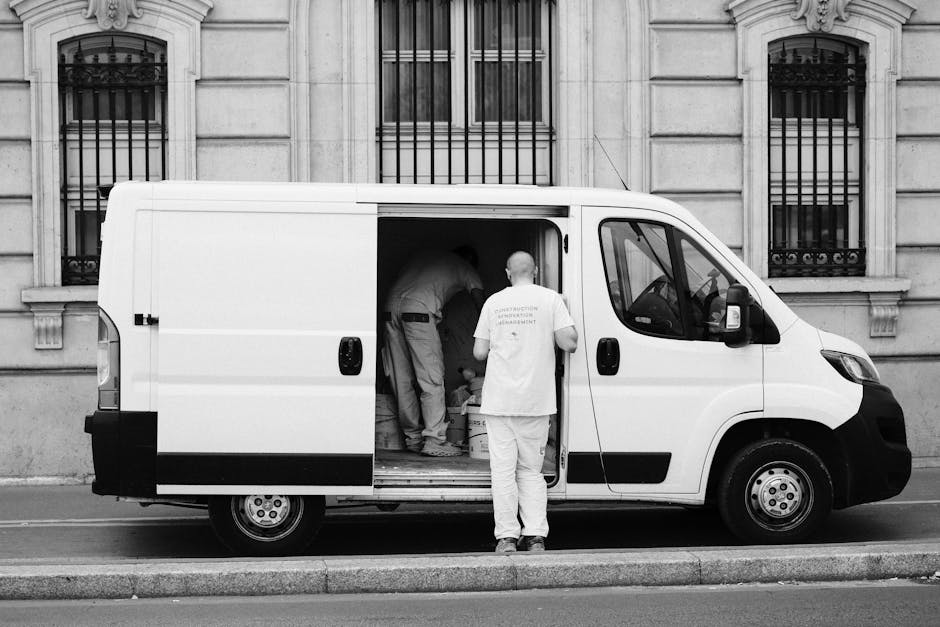 A young man with long hair, wearing a black headband and a dark blue shirt, is sitting in the driver's seat of a white moving van with the door open. He is smiling and giving a thumbs-up gesture, sugg