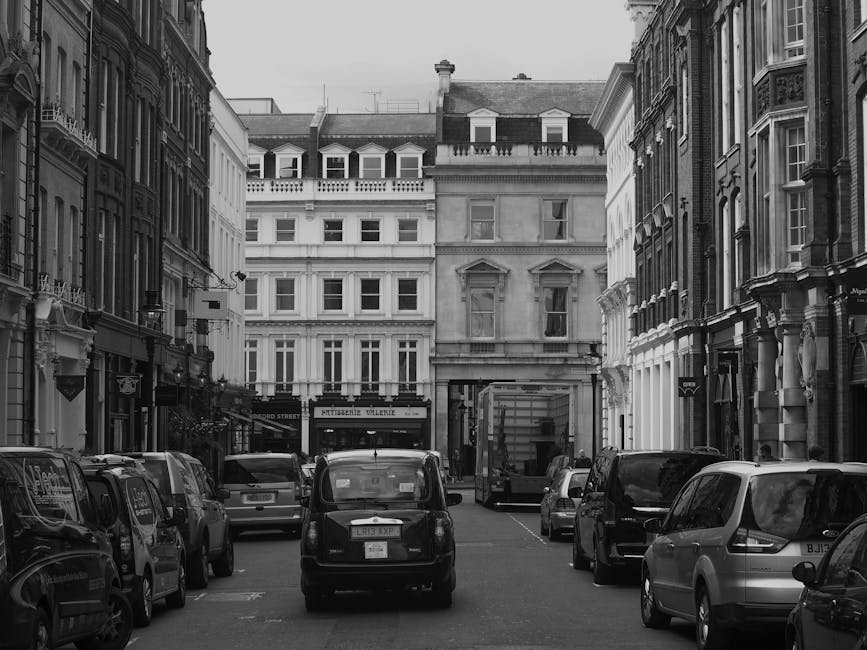 A city street scene showing a narrow road with parked cars on the right side, including black and light-colored vehicles, and a loading area where Self Storage Pimlico is conducting a home relocation.