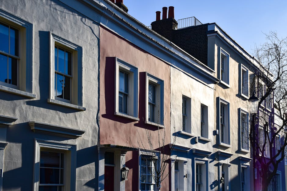 A row of three multi-storey townhouses with pastel-colored facades, including pink, yellow, and blue, situated on a street in Pimlico. The buildings feature white-trimmed sash windows with decorative 